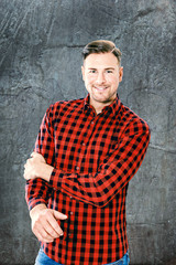 Glücklicher attraktiver bärtiger Mann mit einem strahlenden Lächeln, der in einem bunten roten karierten Hemd  auf seinem Arm fast, vor einem texturierten grauen Hintergrund
