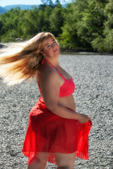 Attraktive plus Größe Frau in bunten roten Outfit posiert im Freien an einem Kiesstrand mit ihren blonden Haaren weht in der Brise mit Blick zurück in die Kamera