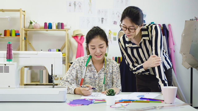 Concept of tailor shop. Attractive teamwork Asian female fashion designers working in workshop. two beautiful coworkers dressmakers women discussing on sketches together in modern bright office.