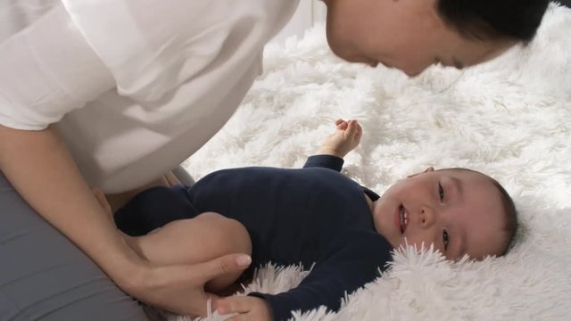 Full shot of happy mixed race baby lying on back, on fluffy white bedspread, trying to turn over, then laughing with delight, while his loving mother is kissing and fondling him