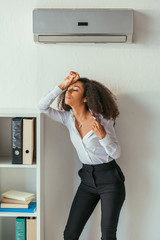 exhausted african american businesswoman holding hand on head while standing under air conditioner in office