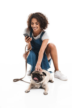 Image of happy african american woman using cellphone and squatting while poising with her pug dog