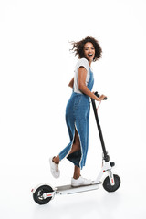 Image of excited african american woman dressed in denim overalls smiling at camera while riding on scooter © Drobot Dean