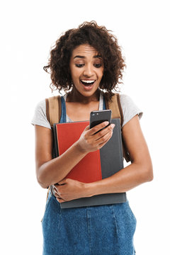 Image of surprised african american woman wearing backpack holding exercise books and cellphone