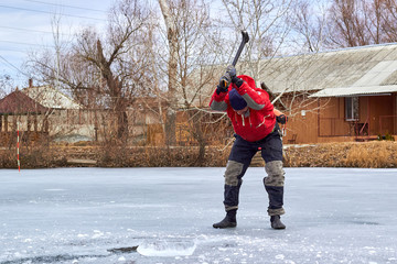 A man in a red jacket pierces the ice on the frozen river with an axe
