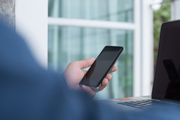 Close up of casual man or freelancer working on laptop computer and holding mobile smart phone with reflection on blank screen, working from home or casual business concept