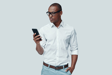 Very busy. Handsome young African man using smart phone while standing against grey background