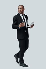 Modern businessman. Full length of handsome young African man in formalwear holding disposable cup and newspaper while standing against grey background