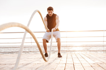 Photo of concentrated man in tracksuit working out with fitness rope on wooden pier at seaside