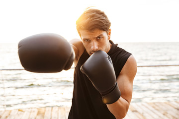 Photo of unshaven man in tracksuit working out in black boxing gloves on wooden pier at seaside