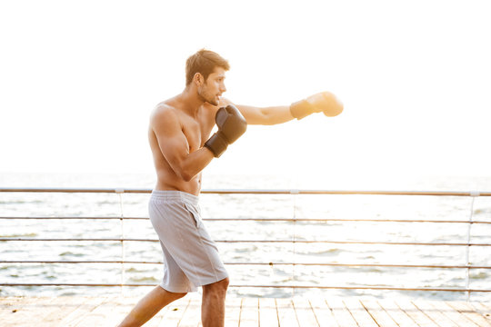 Photo of strong half-naked man working out in black boxing gloves on wooden pier at seaside