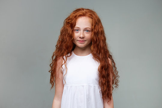 Portrait of cute little girl with ginger hair and freckles, smiles and calming looks at the camera, stands over gray background.