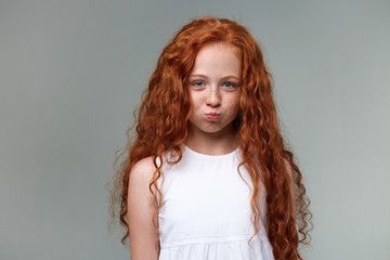 Portrait of cute little girl with ginger hair and freckles, looks at the camera, blows cheeks and...