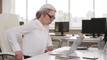 Medium shot of smartly dressed Caucasian businesswoman walking over to her desk in empty office early in morning, sitting down in chair and starting to type email on laptop