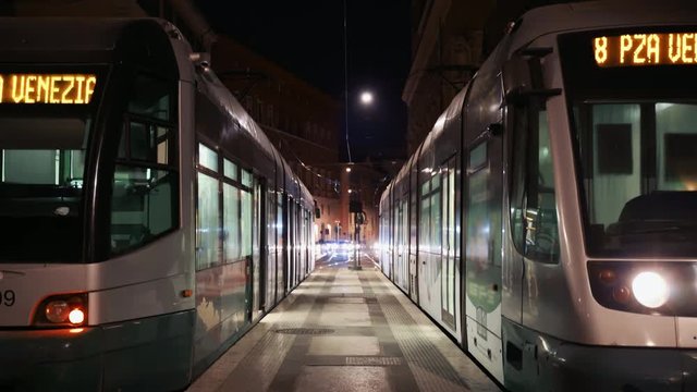 Two trams are on a platform at night in the city of Rome. On the background, cars with fronts are on display. Electric public transport in Italy.