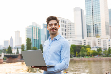 Happy smiling handsome businessman standing outdoors on the street with beautiful sunlight over lake background using laptop computer.