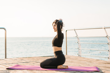 Smiling brunette young woman in a black jumpsuit rests after plank workout on a mat on pier at sunrise.