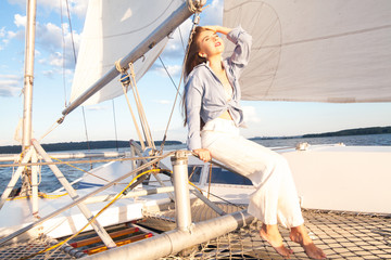 A girl with long hair, a model sits on a yacht, in white pants, a blue shirt, sunglasses, beside a sail, against a blue sky with space for an inscription. The concept of vacation at sea.