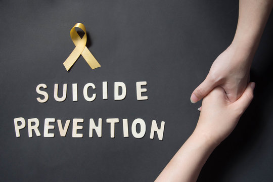 World suicide prevention day - Holding hands for helping and supporting depressed woman with yellow ribbon awareness and SUICIDE PREVENTION wooden word on black background. Mental health care concept.