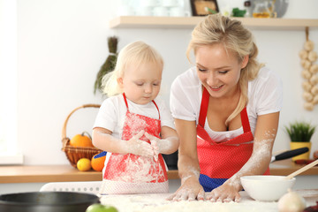 Obraz na płótnie Canvas Little girl and her blonde mom in red aprons playing and laughing while kneading the dough in kitchen. Homemade pastry for bread, pizza or bake cookies. Family fun and cooking concept
