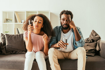 exhausted african american man and woman drinking water from glasses while sitting on couch and suffering from summer heat at home