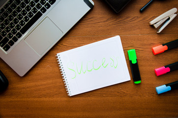 White notepad and a black pen on a wooden desk working place. On the frame is a laptop, purse, colored markers, glasses, smartphone, stapler. Business work success concept, space for text. 