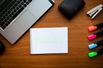 White notepad and a black pen on a wooden desk working place. On the frame is a laptop, purse, colored markers, glasses, smartphone, stapler. Business work success concept, space for text. 