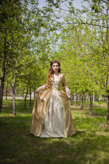 Obraz na płótnie Canvas Beautiful girl in a gold dress against a background of green trees. A woman in a ball gown of the Rococo era.