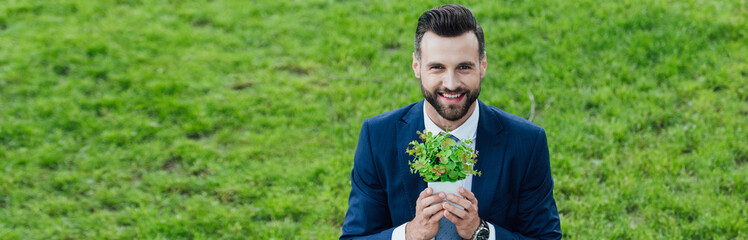 Panoramic shot of young businessman holding flowerpot with plant, smiling and looking at camera