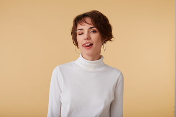 Portrait of playful short haired young woman wears in white golf, winks and looks at the camera, looks so cute and nice, stands over beige wall.