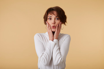 Portrait of amazed short haired young woman in blank t-shirt, looks surprised with wide open eyes, touches cheeks and sens kiss, stands over beige wall. © timtimphoto