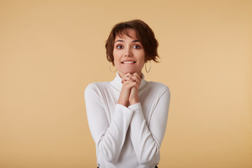 Portrait of beging short haired young woman in blank t-shirt, pitifully looks at the camera, bites lips and cupped hands together, stands over beige wall.