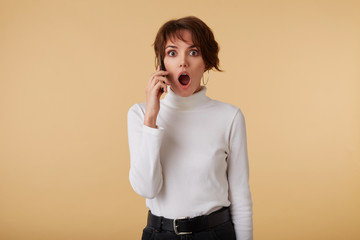 Shocked short haired young woman wears in white golf, holds smartphone and hears unbelievable news,...
