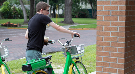 A pale skinned skinny person wearing a black shirt and dark green shorts grabs the handles of a bright lime green bike with yellow rims and a basket about to ride out in the city on a sunny day. - Powered by Adobe