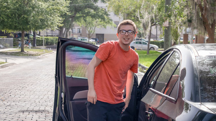 A young pale skinned skinny student leans over his open car door smiling at the camera on the street side in the city with his bright red shirt black glasses shorts and short curly hair on a sunny day