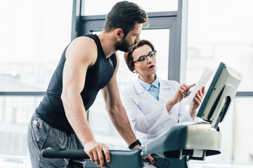 handsome sportsman running on treadmill near doctor with Digital Tablet during endurance test in gym