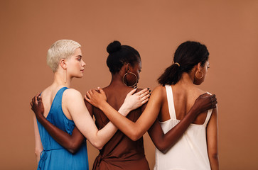 Three diverse women standing against brown background hugging each other