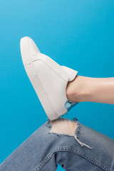Trendy white eco leather sheakers on bent leg knee in ragged jeans over pastel turquoise blue background. Colorful main trend backdrop for your magazine, online store and media. Copy space