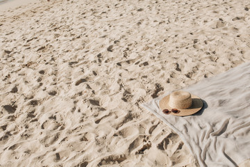 Fototapeta na wymiar Tropical beautiful beach with white sand, foot steps, neutral blanket with straw hat and sunglasses. Relaxing atmosphere. Summer travel or vacation concept. Minimalistic background.