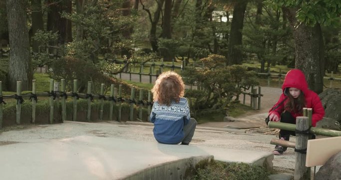 Young boy with curly hair is sitting on the end of a bridge next to a pond in a Japanese ornamental garden in Japan while a young girl in a red jacket squats near by