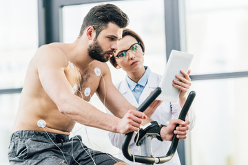 doctor with digital tablet near shirtless sportsman during endurance test in gym