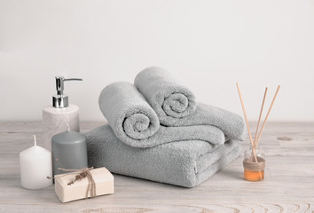 Obraz na płótnie Canvas Rolled and folded gray terry towels with soap, aroma sticks and candles on wooden surface against the white wall. Bathroom items creative composition.