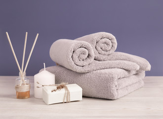 Obraz na płótnie Canvas Rolled and folded gray terry towels with soap, aroma sticks and candles on wooden surface against the lilac wall. Bathroom items creative composition.