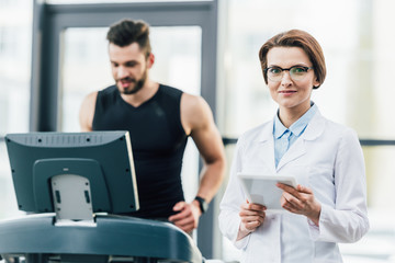 Fototapeta na wymiar sportsman running on treadmill near attractive doctor during endurance test in gym