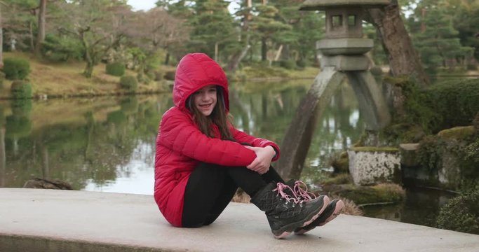 Young girl in a red jacket sitting on a bridge smiling for photos in a Japanese garden in Kanazawa, Japan