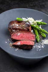 Barbecue dry aged wagyu sirloin beef steak sliced green asparagus and parmesan cheese as closeup on a gray cast iron design plate