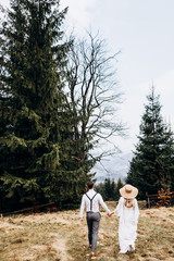 Back view on the Caucasian young just married couple walking hand in hand the countryside meadow with pines. Woman in the long while dress and big hat. Rear. Outdoor.