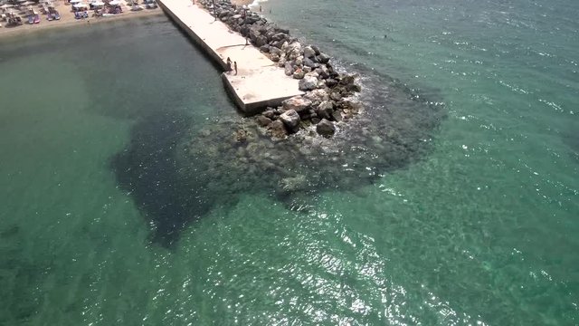 Aerial view of stunning turquoise sea in the summer with pier and rocks that water pops up on them in the area of Agia Paraskevi Halkidiki, Greece, circular movement by drone