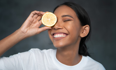 Portrait of beautiful young smiling woman in white t-shirt holding lemon slice in front of eye over concrete background