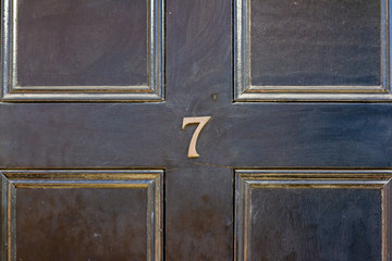 House number seven with the 7 in bronze on a black wooden front door with gold edged carved panels